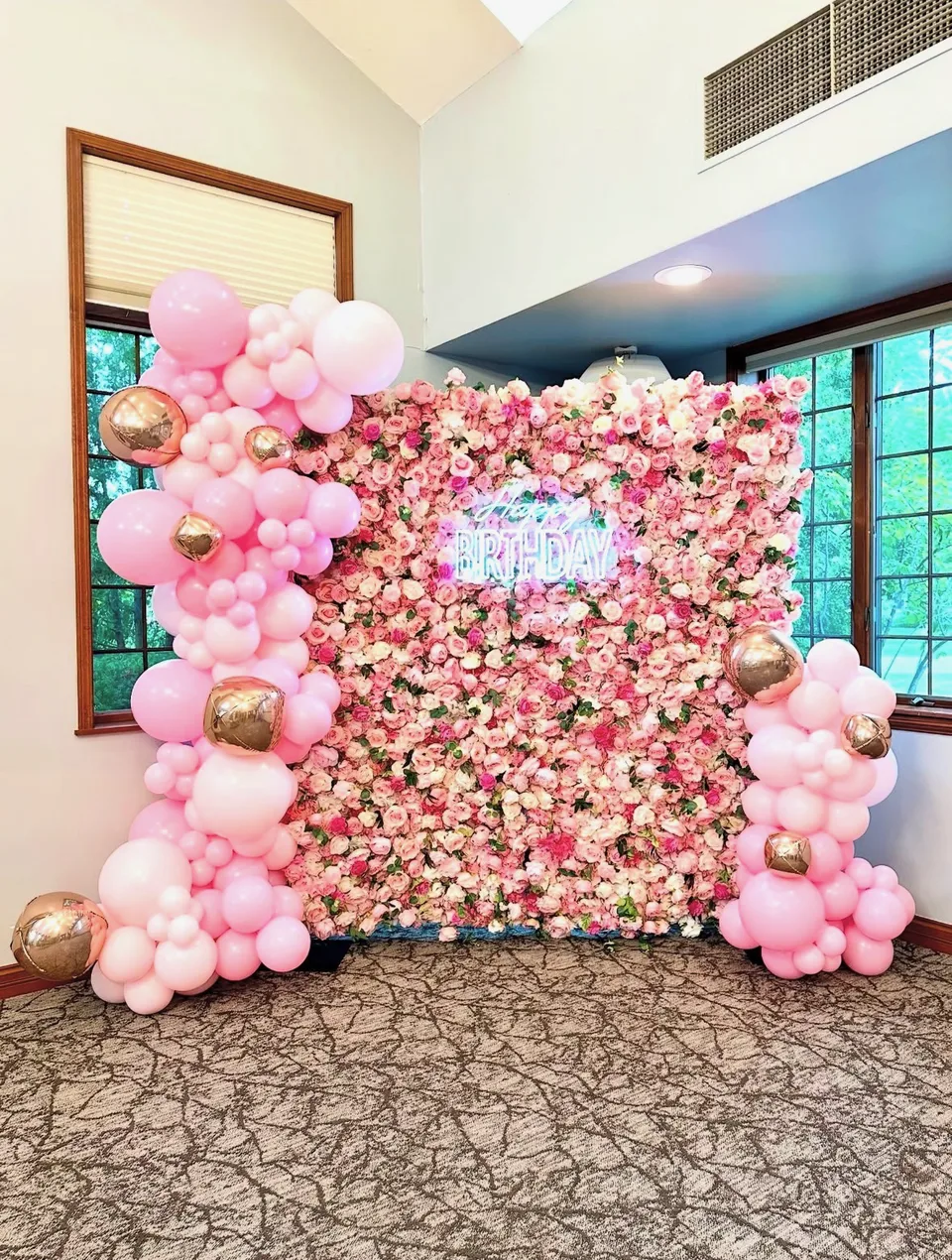 Pink, white and gold balloons framing an indoor flower wall with a birthday neon sign