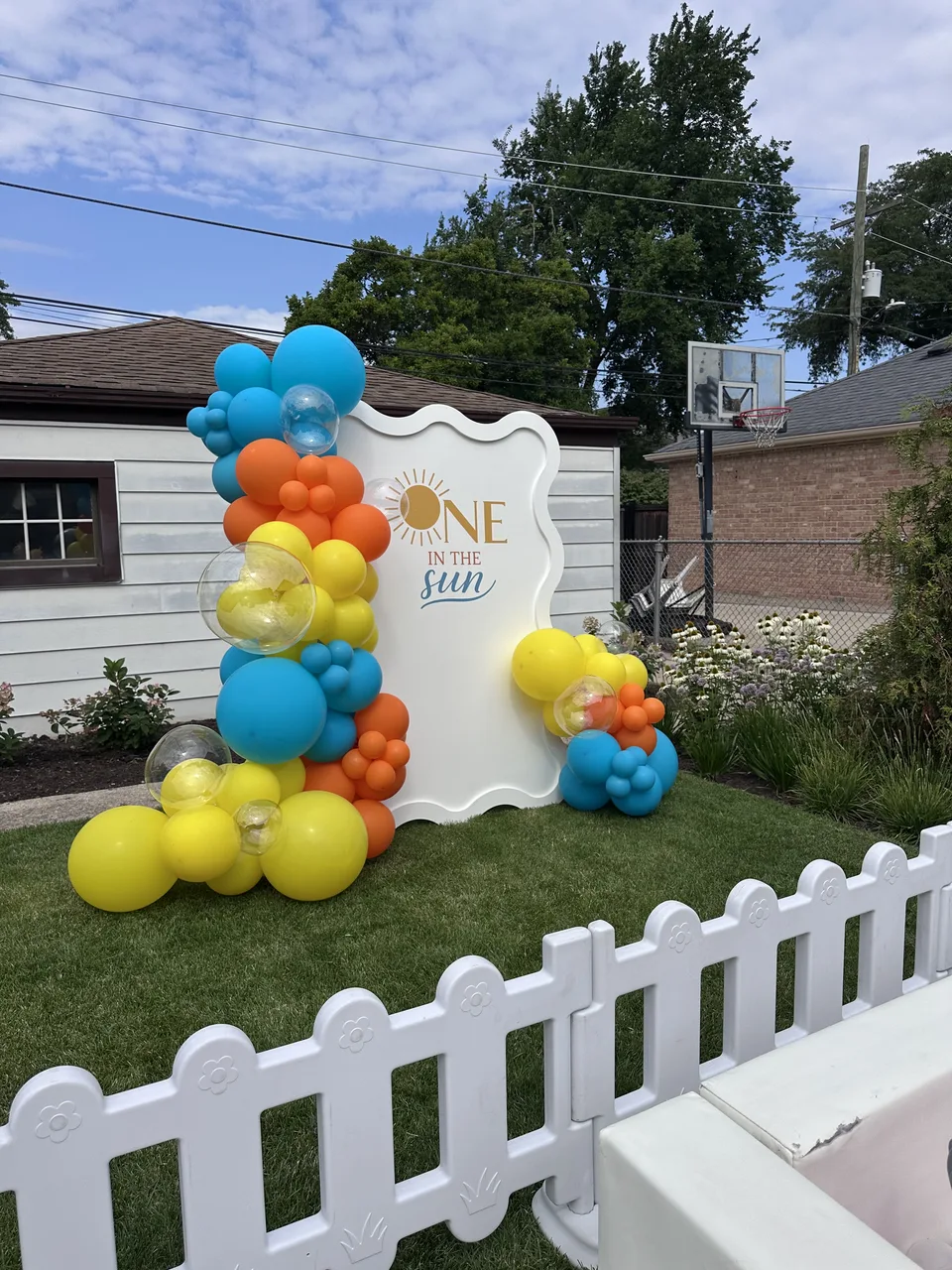 Blue, orange, yellow and white balloons around an outdoor sunshine first-birthday backdrop