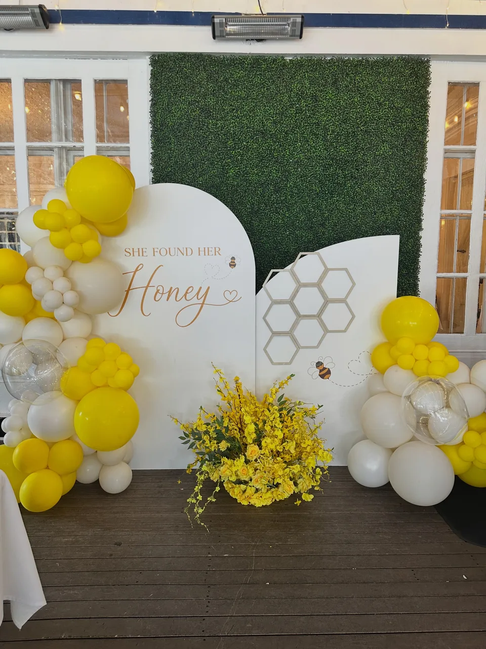 Yellow and white balloons beside honey-themed panels, a hedge wall and yellow flowers